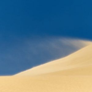Sand dune with sand flying against a blue sky